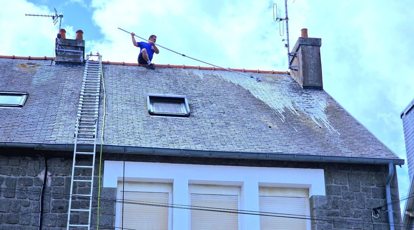 Toiture en ardoise couverte de mousse pendant le nettoyage, technicien assis au faîtage avec une lance